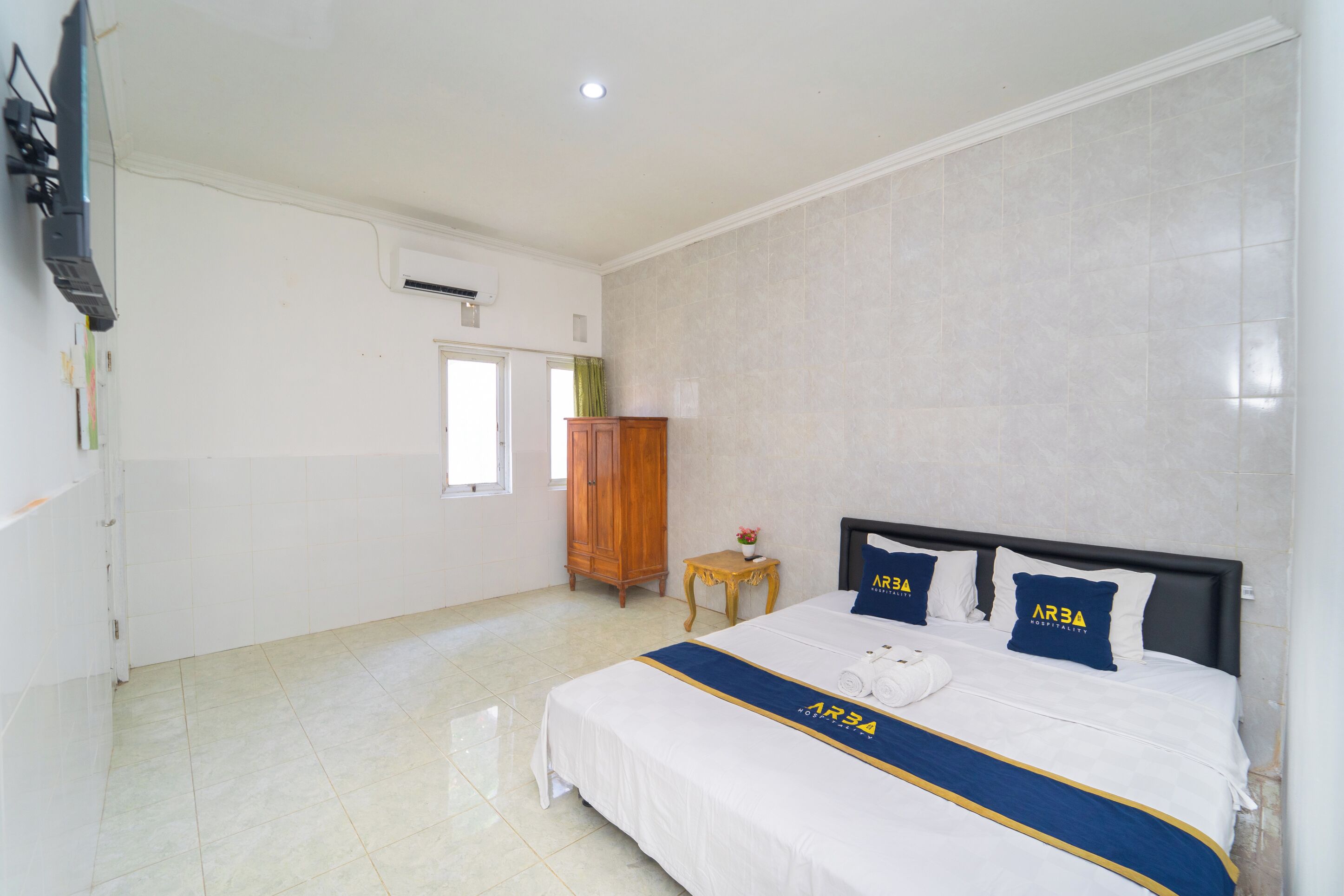 A hotel room with a large bed, a wooden wardrobe, a small table with a vase, and a window with curtains.