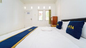 Et hotelværelse med en seng, en blå og gul sengetæppe, en hvid pude med 'AR3 HOSPITALITY' logo, et trænatbord og et vindue med gardin i grøn farve.