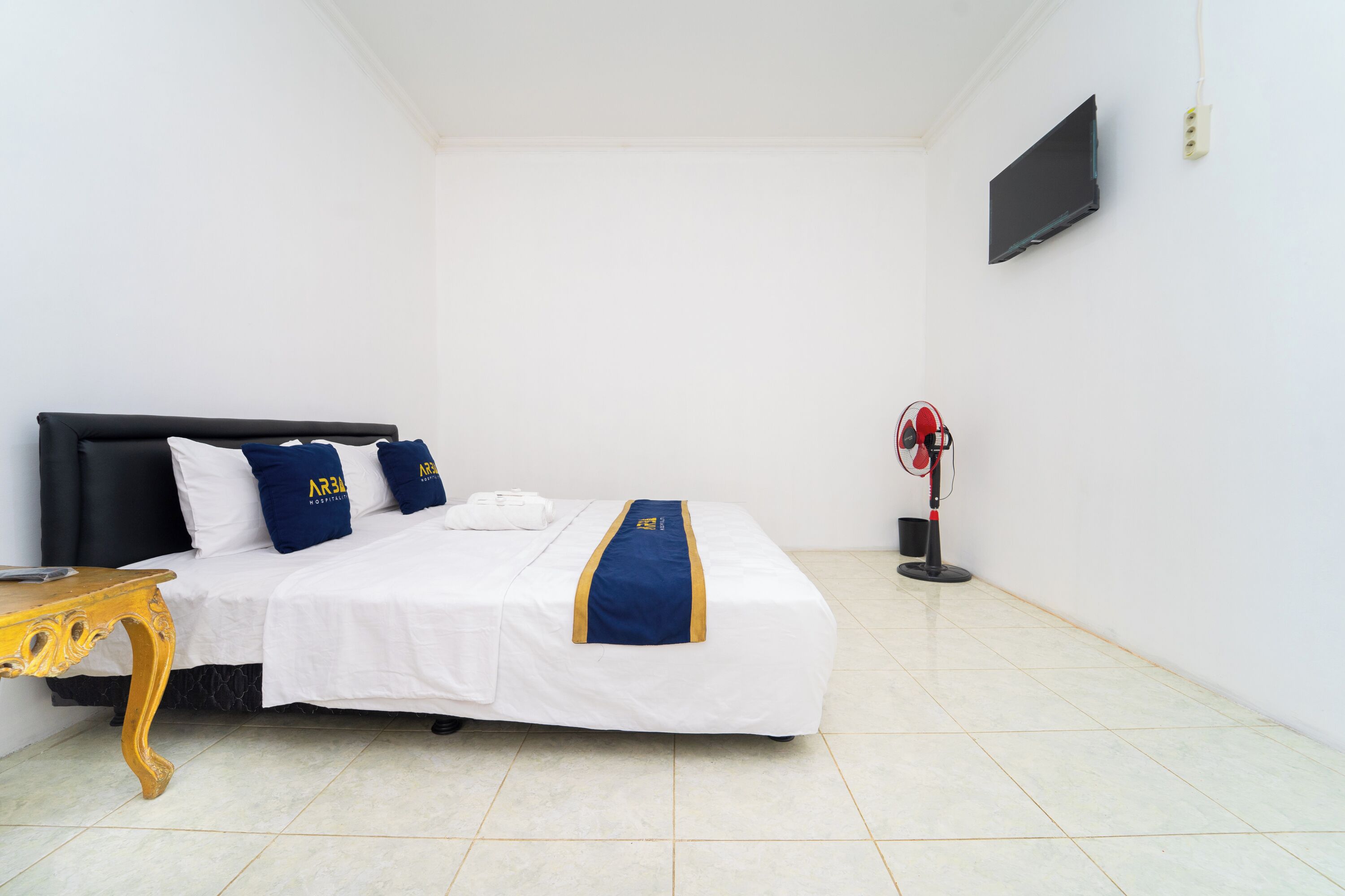 A hotel room with a bed, a wooden nightstand, a red fan, and a TV mounted on the wall.