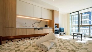 Moderni hotellihuone, jossa on suuri sänky, pieni keittokomero, ruokailutila ja suuri ikkuna, josta avautuu näkymä kaupunkiin.