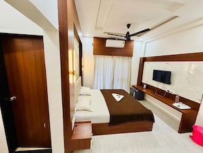 A hotel room with a bed, a desk, a television, and a ceiling fan. - Panchratna Executive Resort (Diveagar)