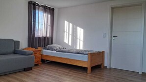 Una habitación con una cama de madera, un sofá gris, una mesita de noche de madera y una puerta blanca.
