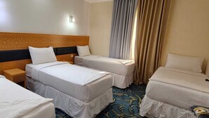 Une chambre d’hôtel avec trois lits simples, une tête de lit en bois, une table de chevet et une fenêtre avec des rideaux.