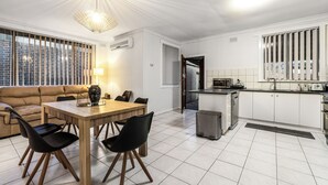 A modern kitchen with white cabinets, a dining area with a wooden table and black chairs, and a large window with blinds. - Generous size 3BR 1.5Bath Every BD has AC (Oakleigh South)