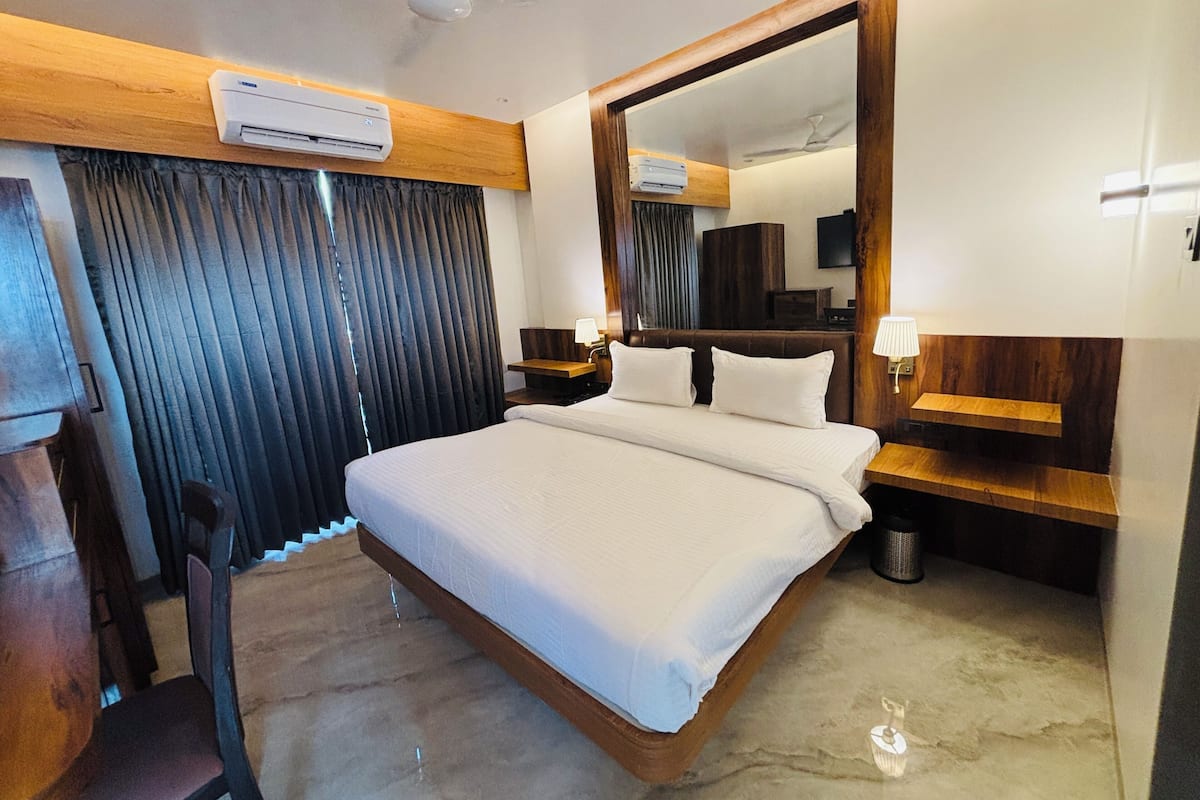 A hotel room with a large bed, a wooden headboard, a mirror, a desk with a lamp, and an air conditioning unit.