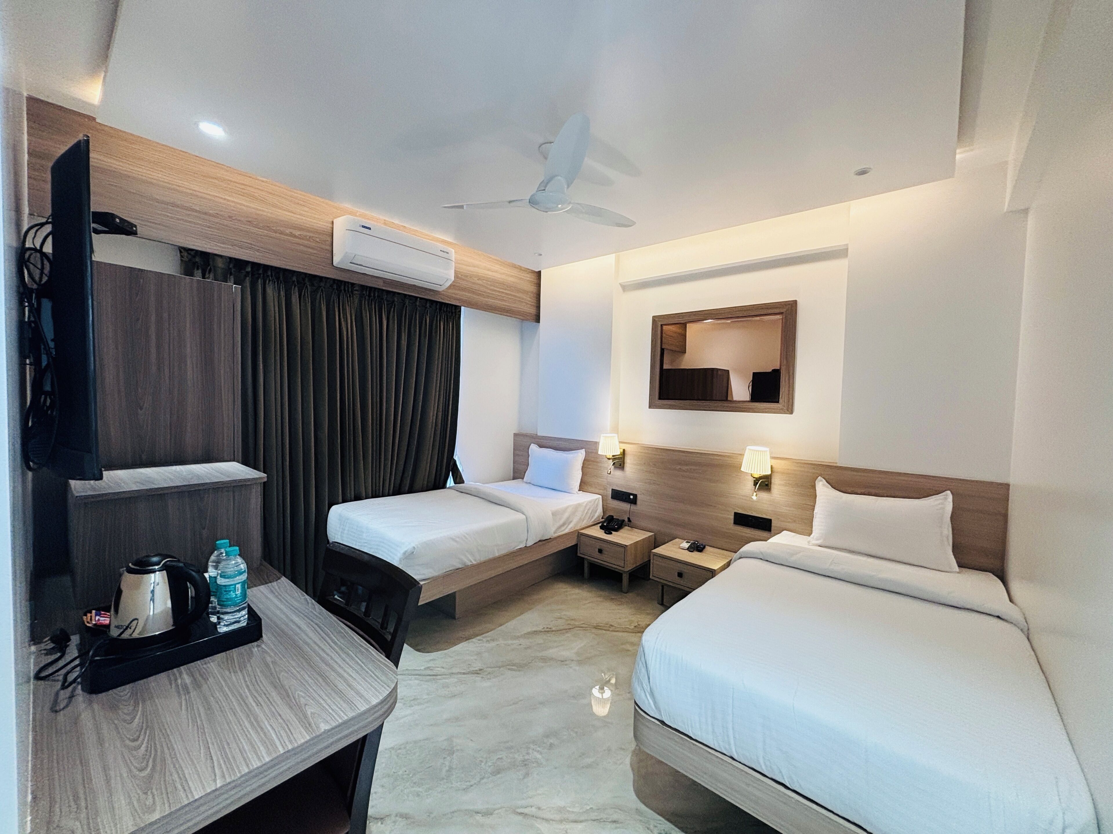Habitación de hotel con dos camas, un escritorio con tetera y botella de agua, aire acondicionado, ventilador de techo y un espejo en la pared.