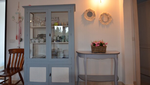 A dining area with a blue cabinet containing glassware and dishes, a wooden chair, and a small table with a basket of flowers. - Fsl-quas01 in San Lorenzo (Budoni)