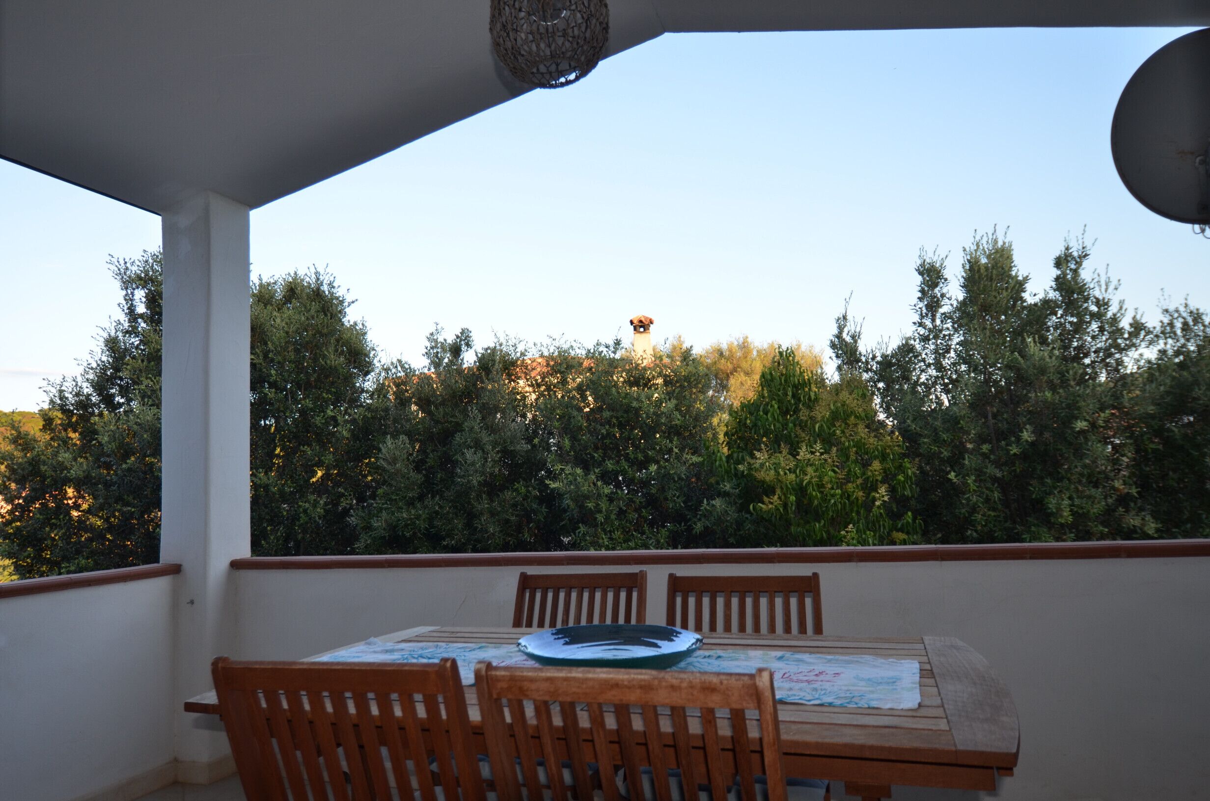 A balcony with a table and chairs, overlooking a garden with trees and a distant tower.