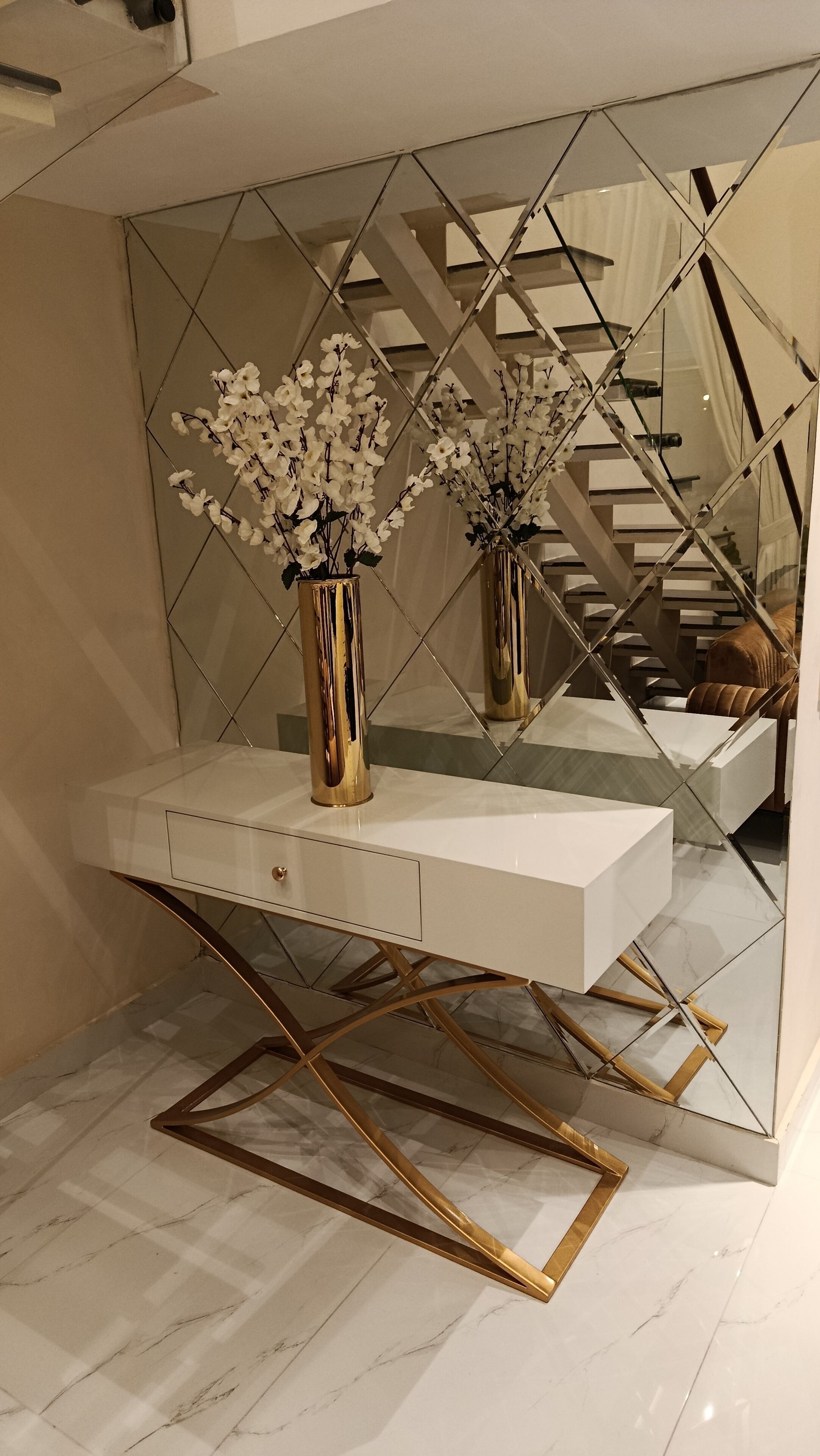 Ein modernes Interieur mit einem weißen Konsolentisch, einer goldrahmigen Glasplatte und zwei goldenen Vasen mit weißen Blumen.