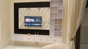 A modern living room with a flat-screen TV mounted on a marble-patterned wall, a white entertainment unit with drawers, and a glass-fronted shelving unit with decorative items.