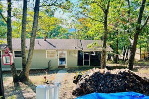 A single-story house with a front porch, a garage, and a yard with trees and a pile of firewood. - Charming Mid-Century Cabin - Warm, Cozy & Scenic (Fort Loudon)