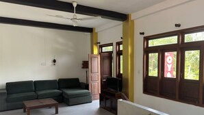 A living room with a sectional sofa, a coffee table, a television, and a window with a view of greenery. - Eros Hotels Green house (Unawatuna)