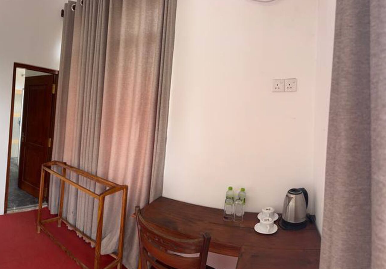 A room with a wooden table, two chairs, a red carpet, a window with curtains, a kettle, and two bottles on the table.