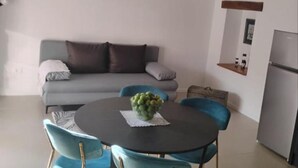 Un salon moderne avec un canapé gris, une table ronde entourée de chaises, un petit coin cuisine doté d’un réfrigérateur et un luminaire fixé au mur.
