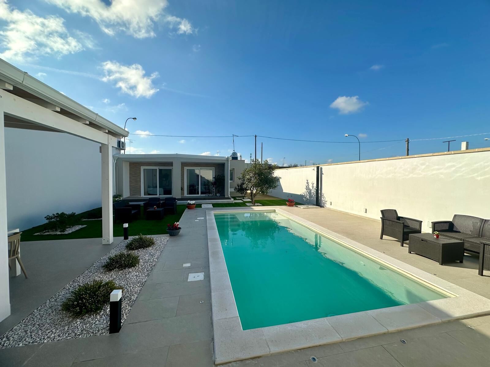 Un espace piscine extérieur moderne, doté d’une piscine à l’eau d’un bleu limpide, entourée d’un mur blanc et d’un petit jardin.