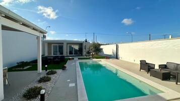 Un espace piscine extérieur moderne, doté d’une piscine à l’eau d’un bleu limpide, entourée d’un mur blanc et d’un petit jardin.