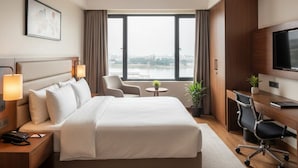A hotel room with a bed, bedside lamps, a desk, a chair, a TV, and a view of a body of water. - Hotel Anand Regency Riverfront (Dhule)