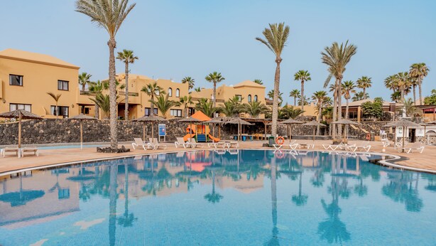 A swimming pool with lounge chairs and palm trees, surrounded by buildings with balconies and a play area. - Casa  Emma (isla de Fuerteventura)