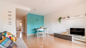 A modern living room with a teal accent wall, a dining area with white chairs, a flat-screen TV, and a sofa with colorful cushions. - Casa  Emma (isla de Fuerteventura)