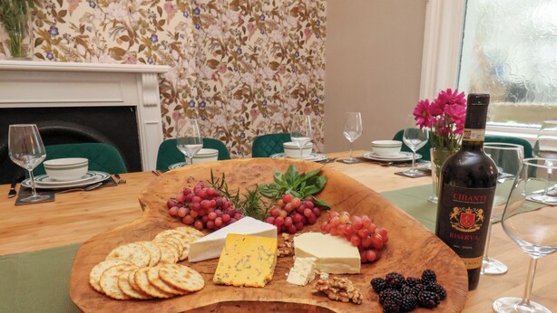 Een houten plank met kaas, druiven en crackers op een tafel in een eetruimte, met groene stoelen en behang met bloemen.