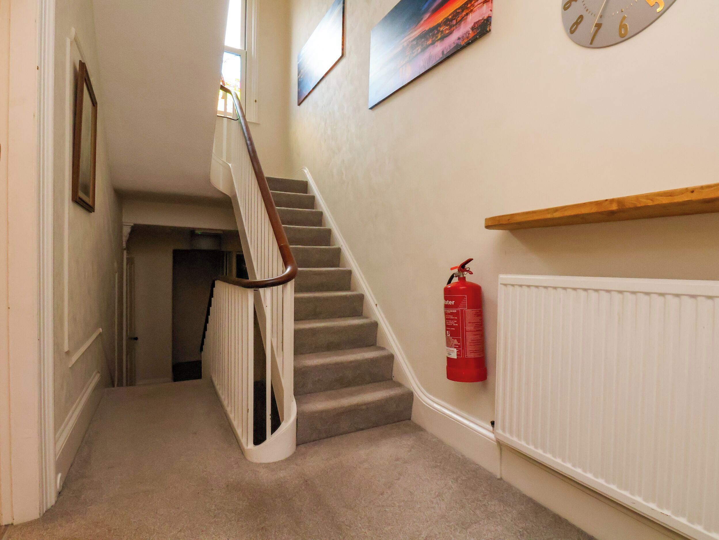 A carpeted staircase with a wooden handrail, a fire extinguisher mounted on the wall, and a clock.
