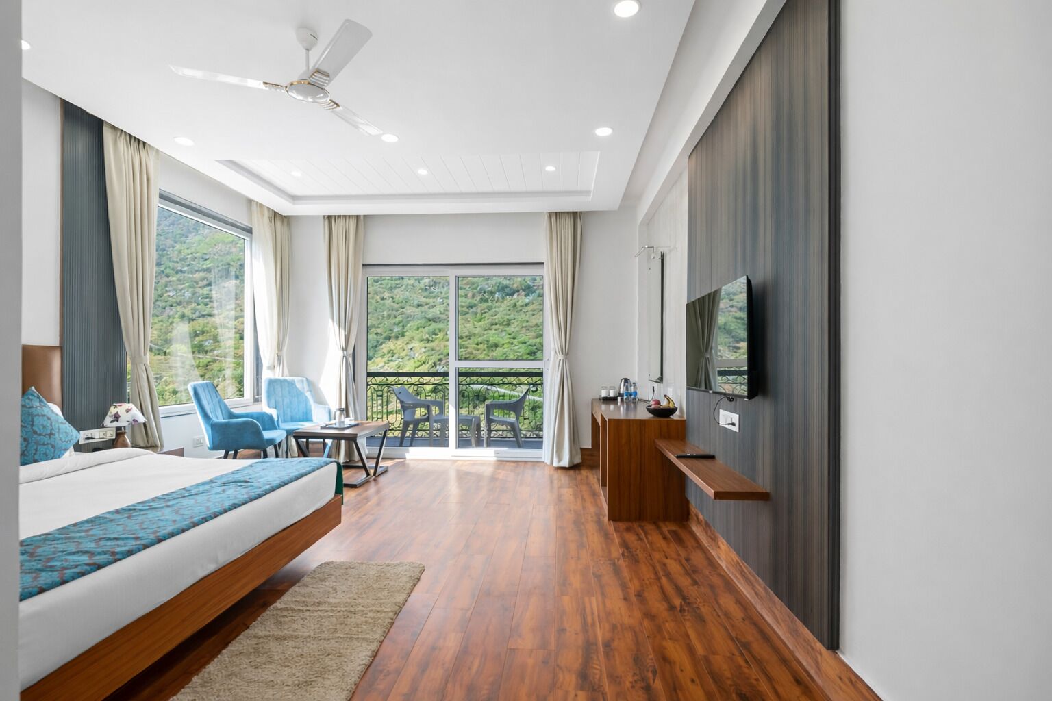 A modern hotel room with a bed, a desk, a chair, a TV, and a balcony with a view of greenery.