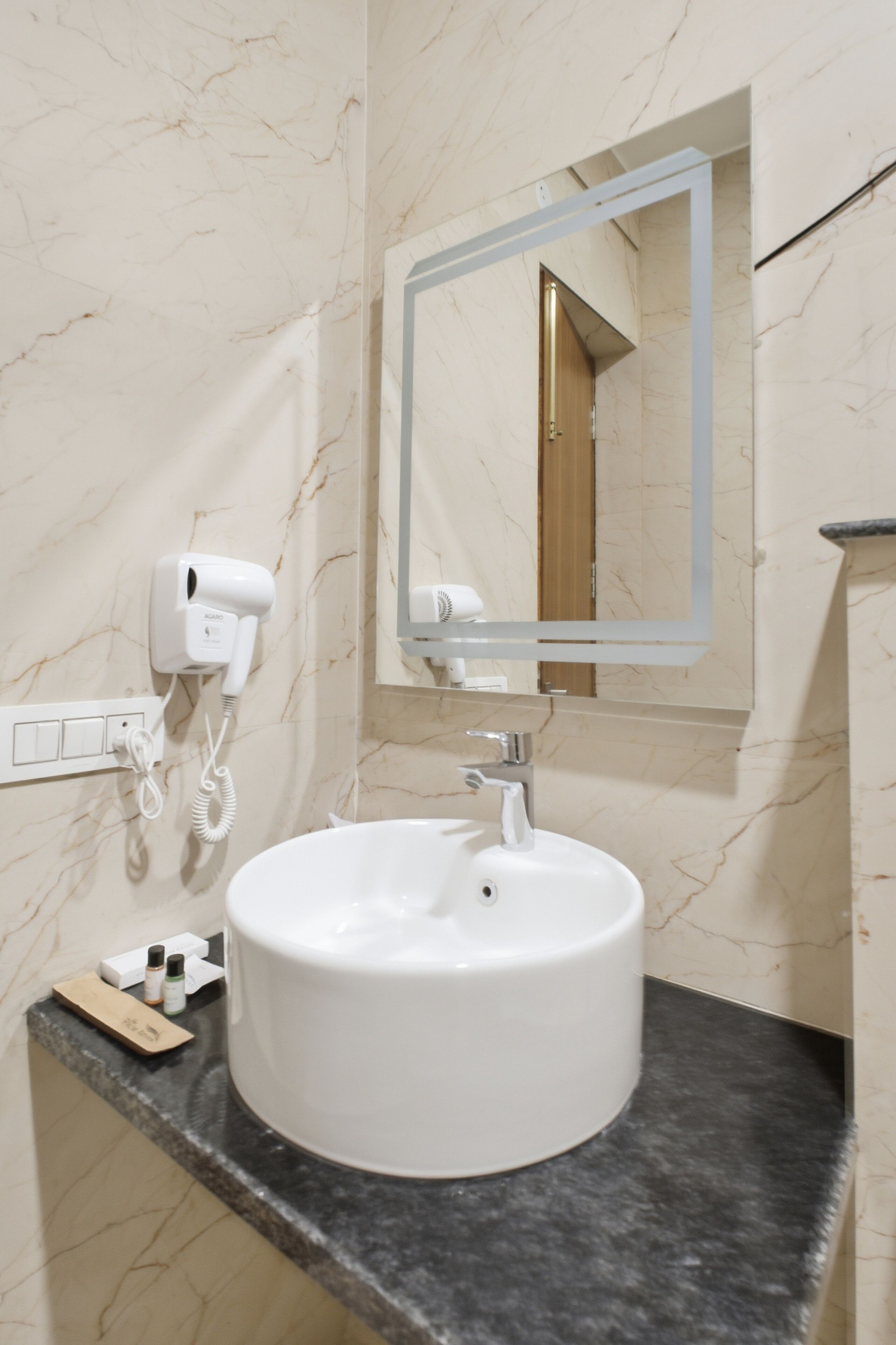A bathroom with a marble wall, a white sink, a mirror, and a hairdryer mounted on the wall.