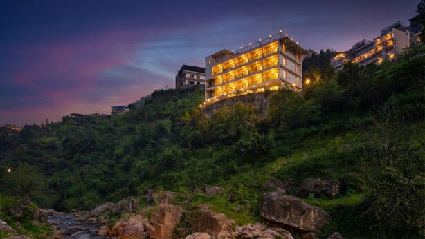A multi-story hotel with balconies, situated on a hillside overlooking a flowing river. - Palm Aryan Pause by the Stream Resort (Dhanaulti)