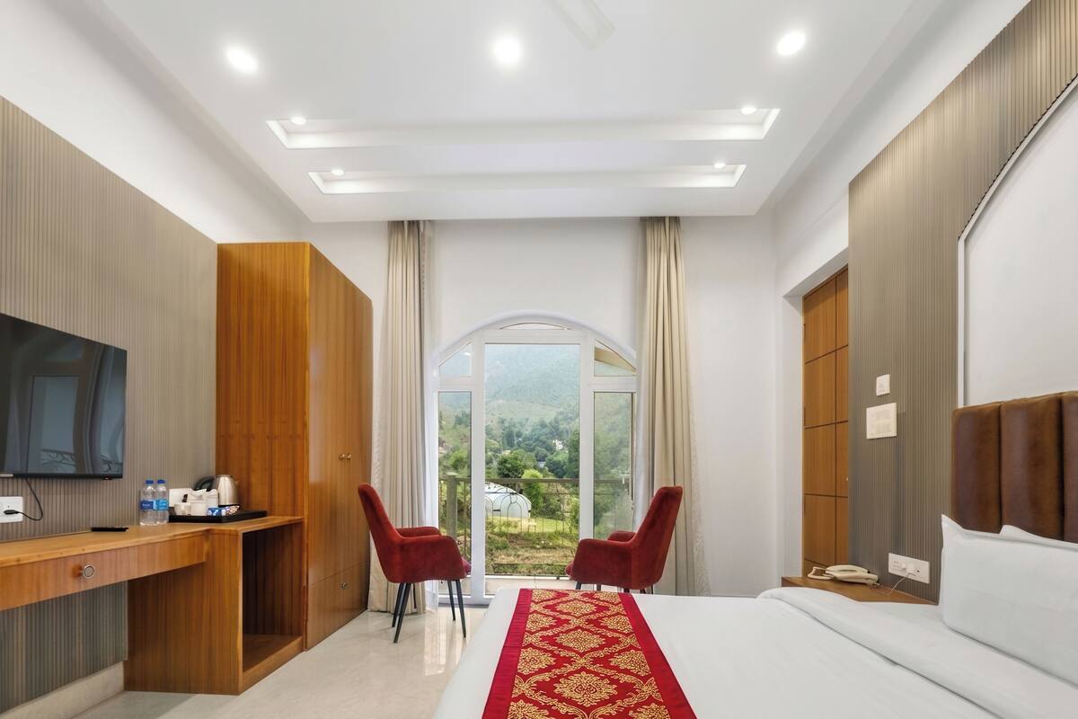 Habitación de hotel con cama, escritorio con televisor, dos sillas rojas, armario de madera y un ventanal con vistas.