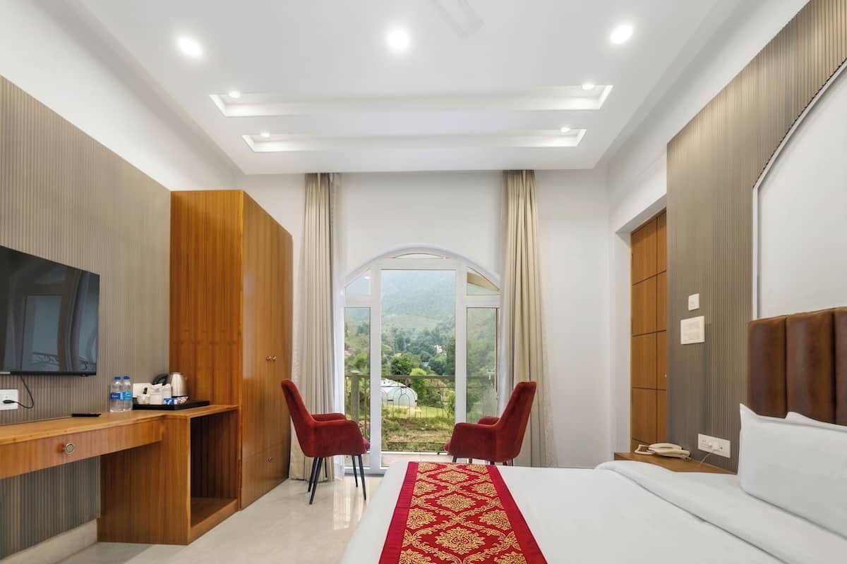 Habitación de hotel con cama, escritorio con televisor, dos sillas rojas, armario de madera y un ventanal con vistas.