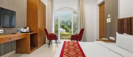 Une chambre d’hôtel avec un lit, un bureau avec une télévision, deux chaises rouges, une armoire en bois et une grande fenêtre donnant sur un beau paysage.