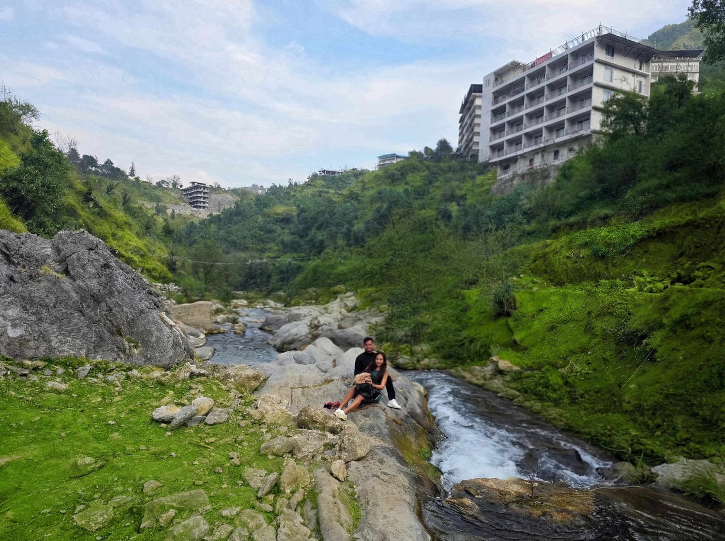 A person sitting on rocks by a flowing river, surrounded by lush greenery and a building in the background.