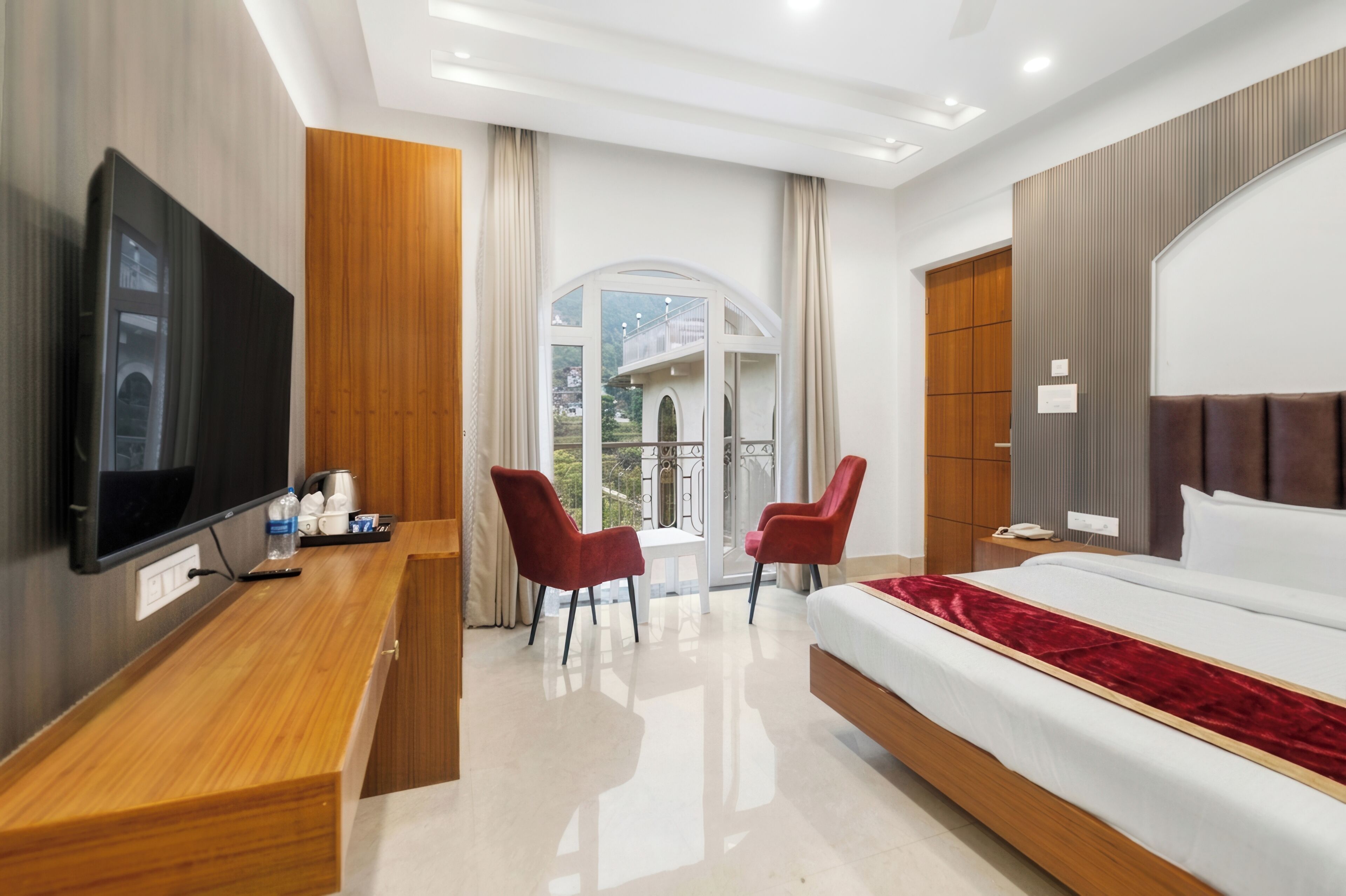 A modern hotel room with a bed, TV, desk, chairs, and a balcony with a view.