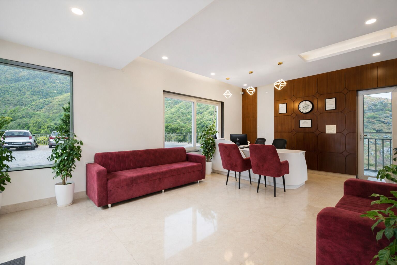 Un espace d’accueil moderne avec un canapé rouge, deux fauteuils, un bureau et un ordinateur. De grandes fenêtres offrent une vue sur un parking et sur des espaces verts.