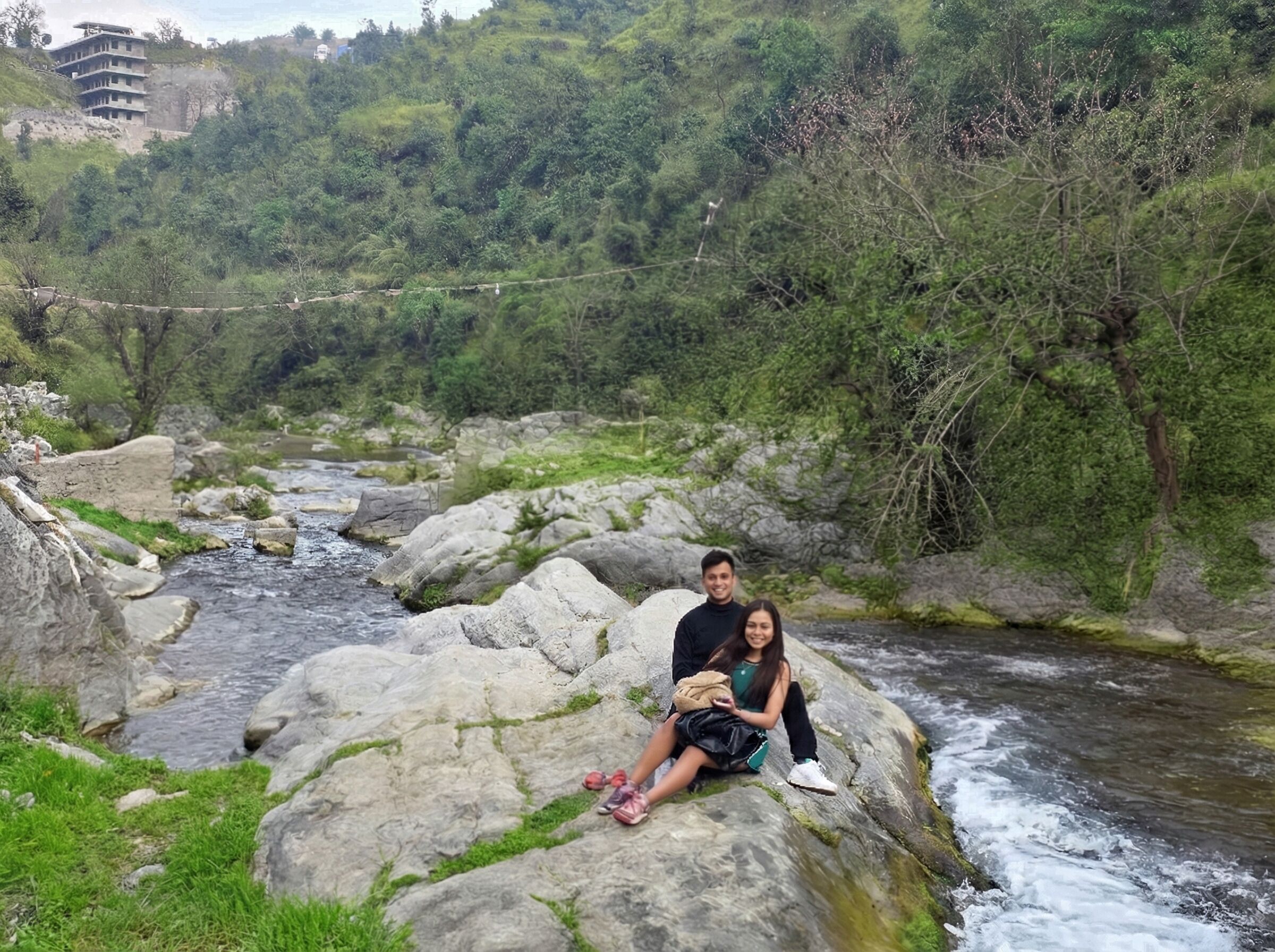 Una pareja sentada sobre una roca grande junto a un río que fluye, rodeada de exuberante vegetación y un edificio al fondo.