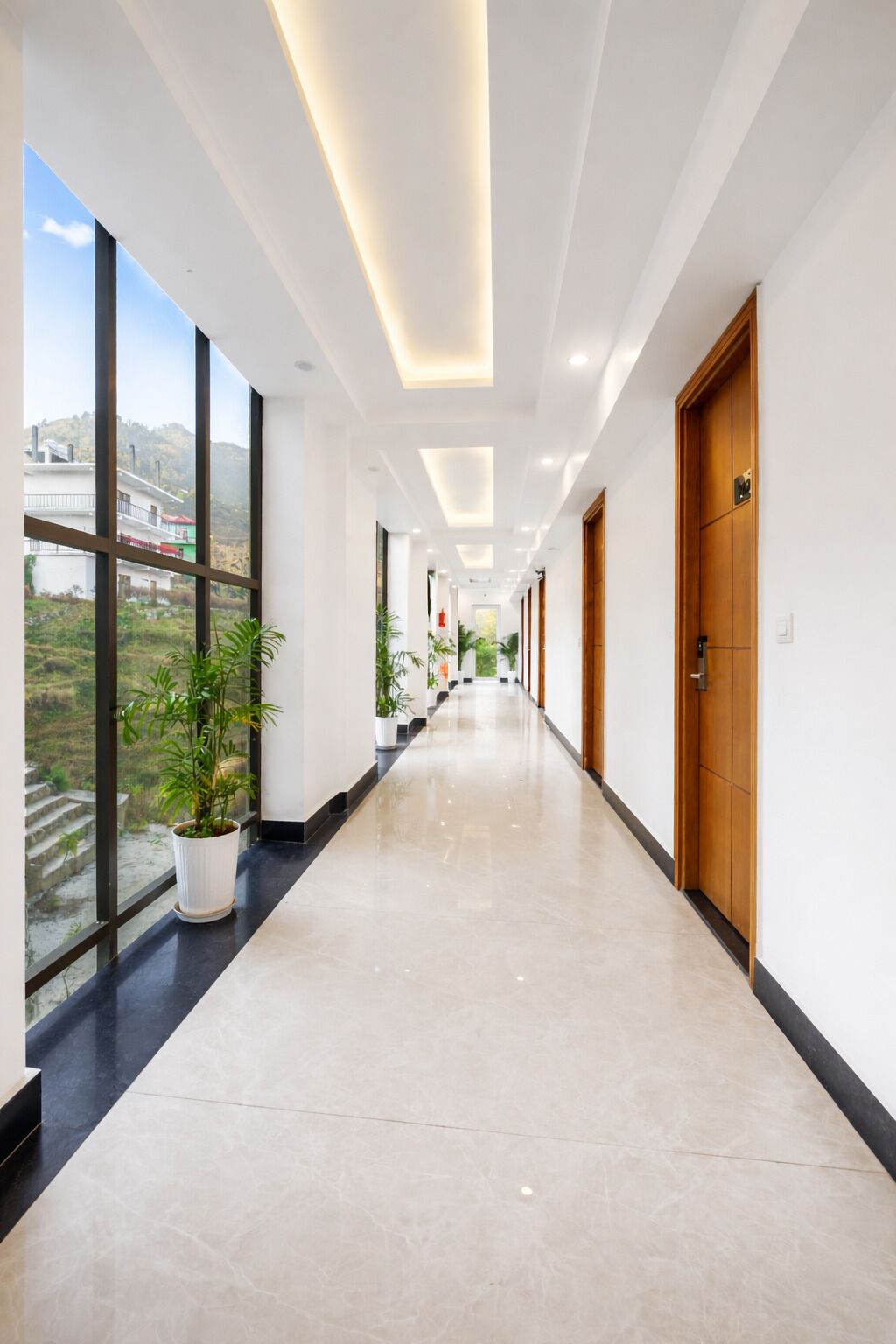 Un largo pasillo bien iluminado con amplias ventanas a un lado, plantas en macetas y una puerta de madera al final.