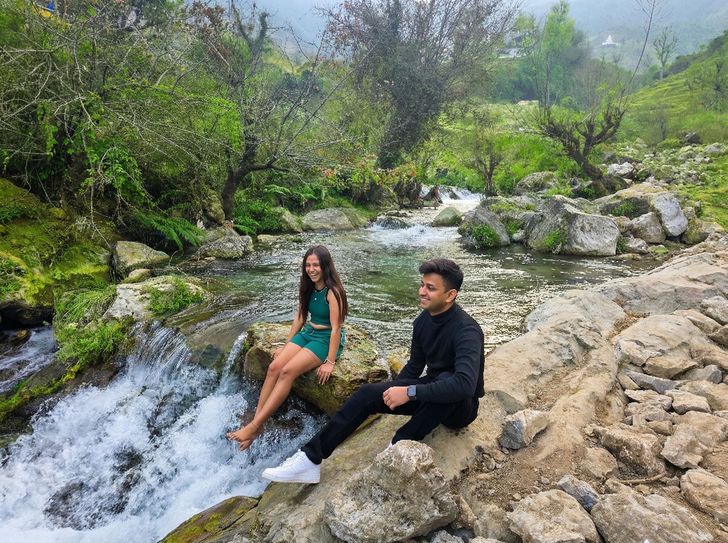 Two people sitting on rocks by a flowing stream in a lush, green environment.