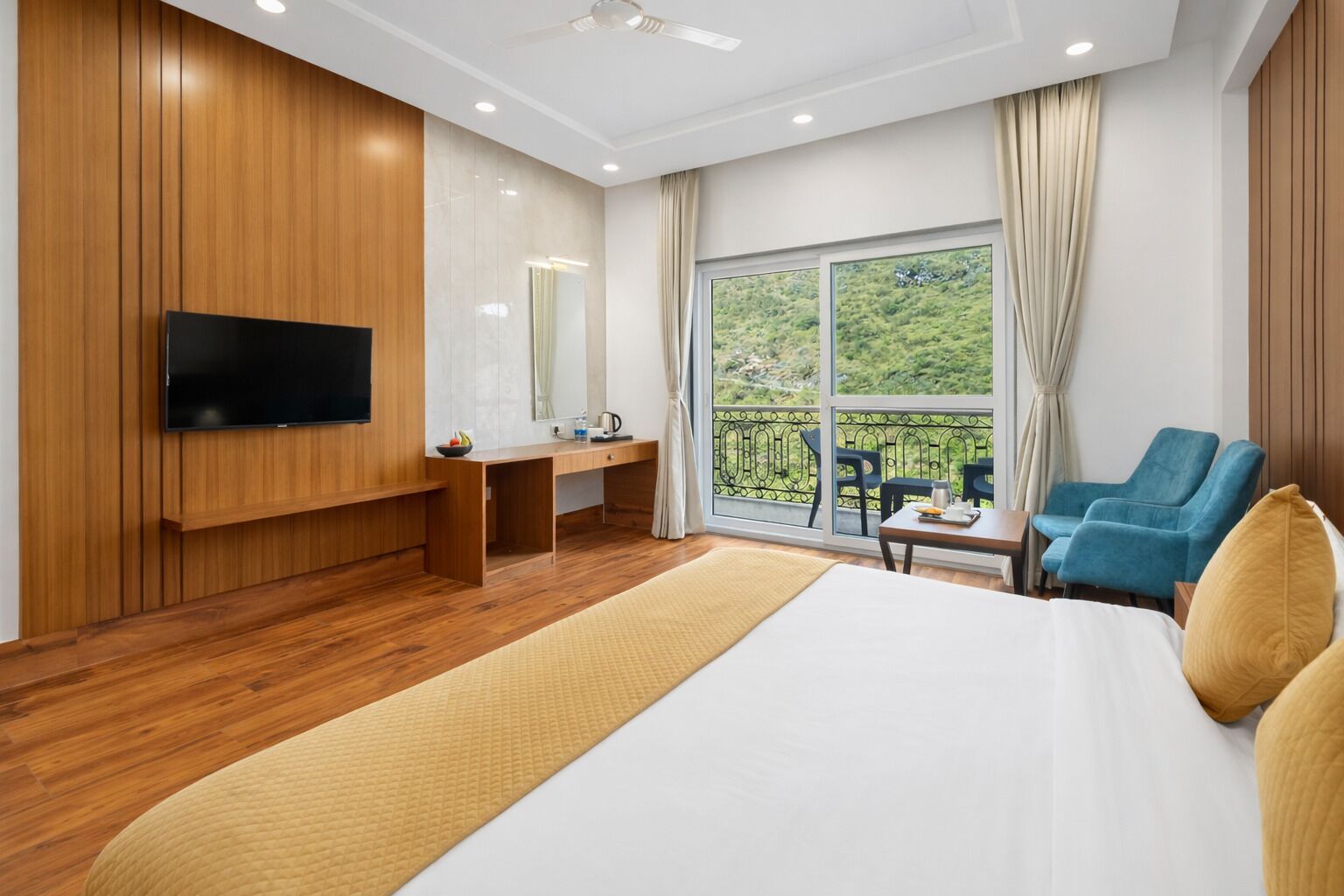 Une chambre d’hôtel moderne avec un plancher en bois, un grand lit, un téléviseur fixé au mur, un bureau avec une chaise et un balcon offrant une vue sur la verdure.