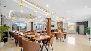 A spacious dining area with multiple tables and chairs, a high ceiling with recessed lighting, and large windows offering a view of the outdoors. - Palm Aryan Pause by the Stream Resort (Dhanaulti)