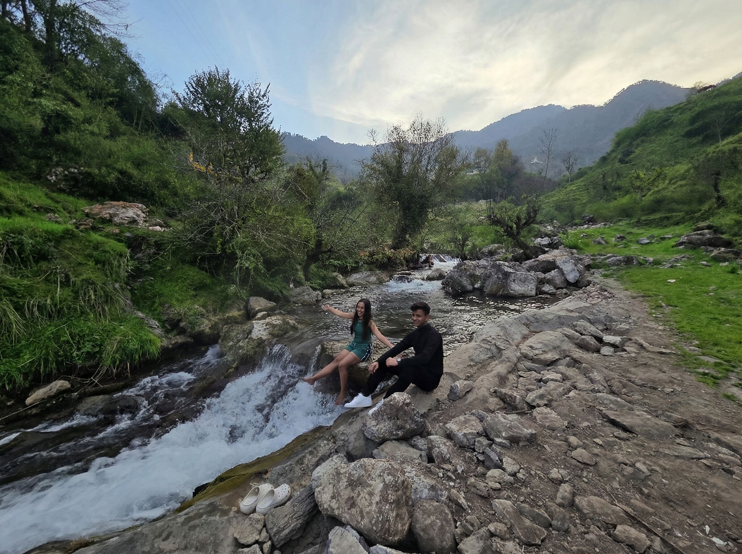 Two people sitting on rocks by a flowing stream in a mountainous area.