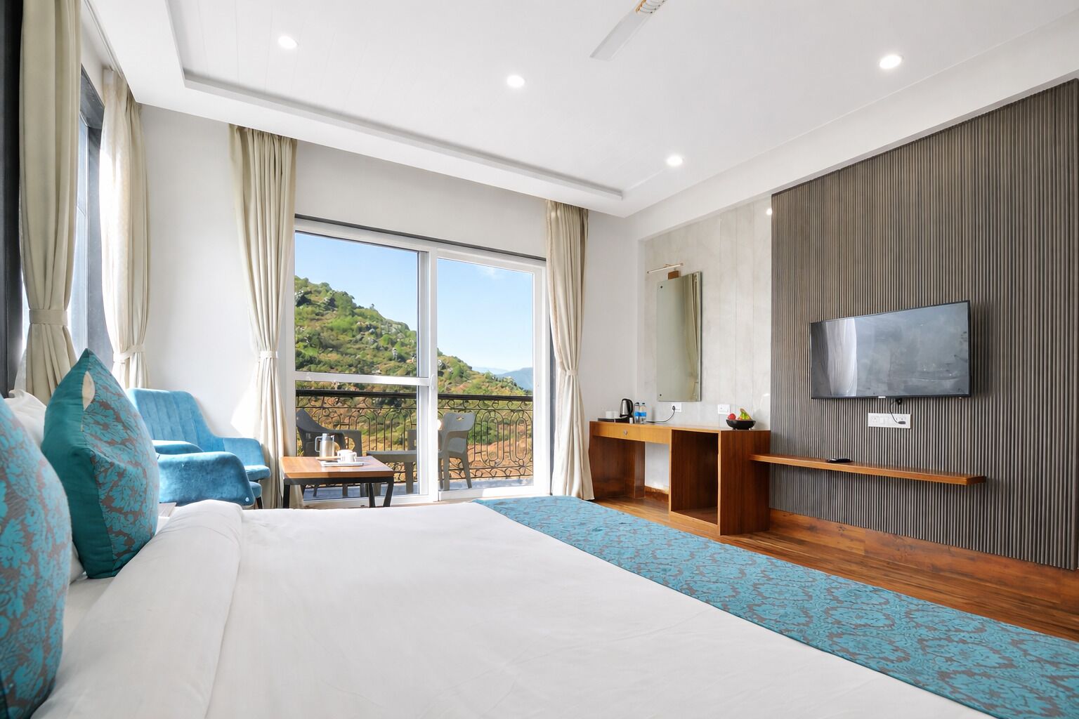 A modern hotel room with a large bed, a desk, a TV, and a balcony with a scenic view.