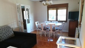 Un salon avec un canapé noir, une table basse en verre, une table à manger avec des chaises blanches et un téléviseur posé sur un meuble en bois.
