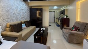 A modern living room with a sofa, a coffee table, and a TV. There is a kitchen area with cabinets and a refrigerator. - Room With AirPurifier ,Recliner Near kolkata Airprot, CC2 and Convention Center (Kolkata)
