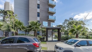 A modern building with a glass entrance, surrounded by palm trees and parked cars. - Studio in Condominium w/ Gym and Laundry Eco0304 (Curitiba)