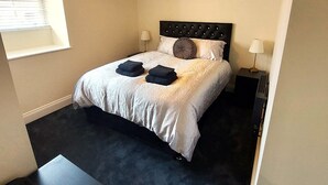 En pænt redt seng med en knaploft, to sengeborde og et lille bord i et rum med beige vægge og et mørkt tæppe.