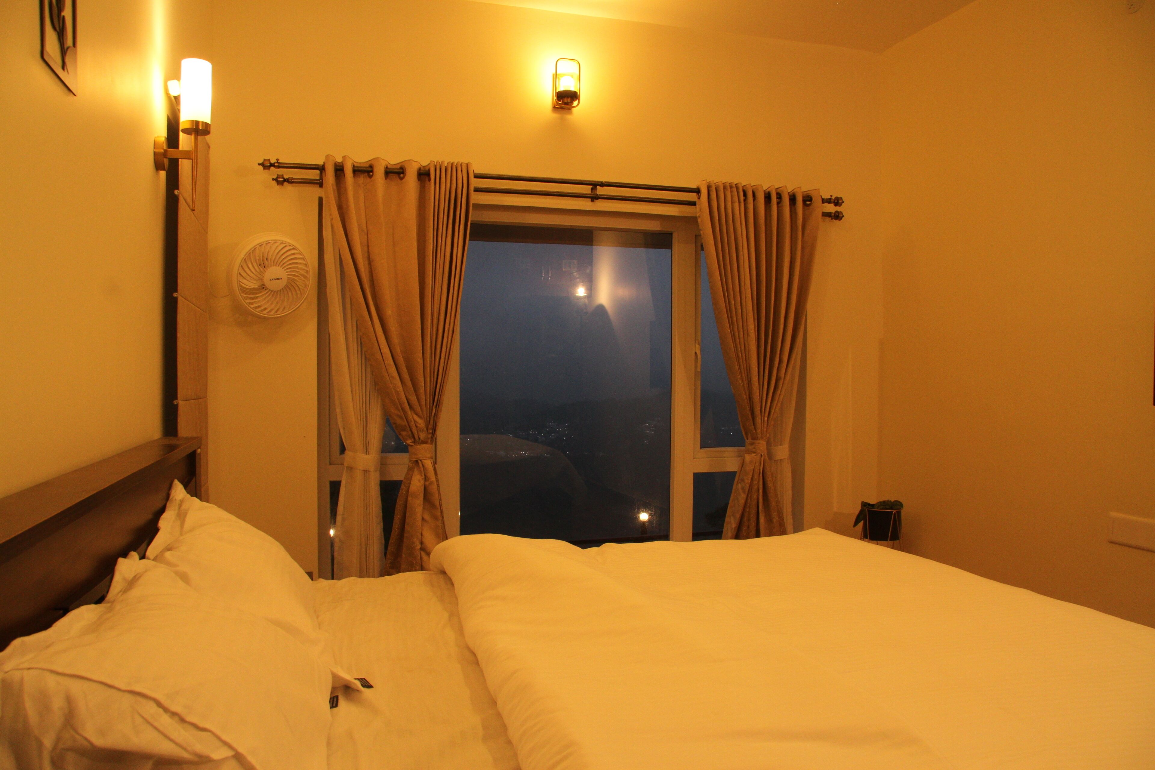 Une chambre d’hôtel avec un lit, une fenêtre avec des rideaux, un ventilateur fixé au mur et un luminaire au-dessus de la fenêtre.