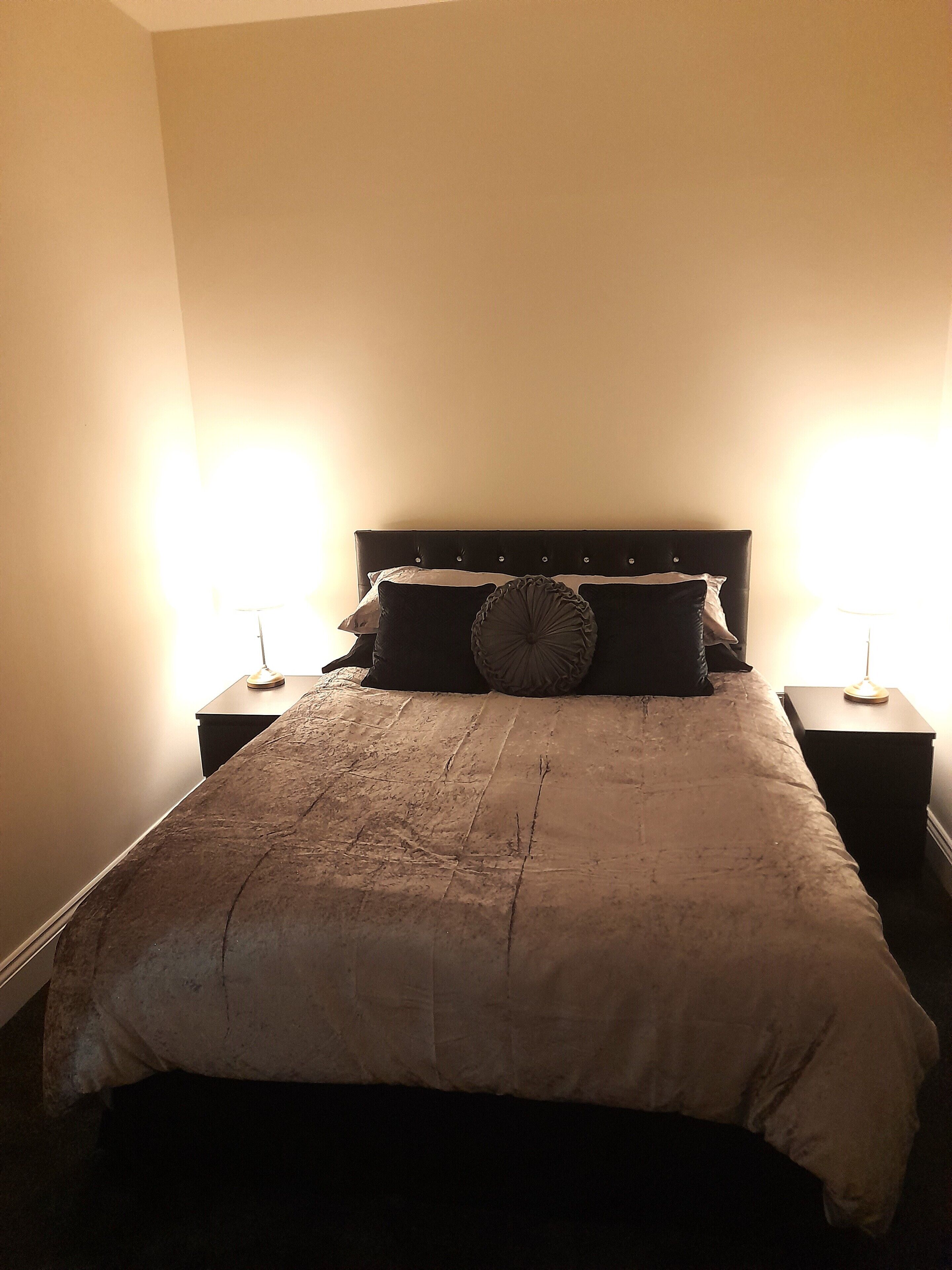 Una cama con cabecera oscura, dos mesitas de noche con lámparas y una pared lisa.