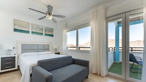 Une chambre d’hôtel moderne, équipée d’un lit, d’un canapé gris, d’un ventilateur de plafond et d’un balcon offrant une vue sur le désert.