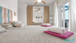 Une chambre avec deux lits, un ventilateur de plafond, une grande fenêtre avec des rideaux et une vue sur la mer.