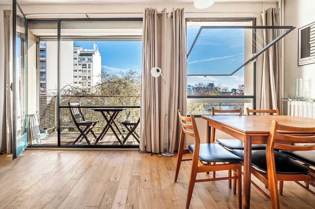 A room with a dining table and chairs, a balcony with a view of buildings, and a large window with curtains.
